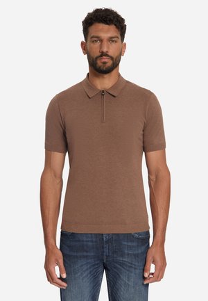 Man draagt een bruine polo met korte mouwen en een rits aan de voorkant, en een blauwe spijkerbroek, staand tegen een effen witte achtergrond.