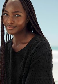 Sort strikket sweater med åbent vævemønster, rund halsudskæring og tekstureret finish. Båret af en person med langt fletninger, foran en strandbaggrund.