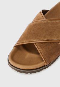 Ruskea mokkanahkainen sandaali, jossa leveät ristikkäiset remmit ja pehmustettu jalkinepohja, teksturoidulla kumipohjalla.