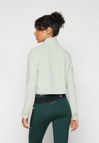 Hellgrüne cropped Jacke mit hohem Kragen und langen Ärmeln, aus weichem Stoff gefertigt. Getragen über dunkelgrünen Leggings mit schwarzen Akzenten.