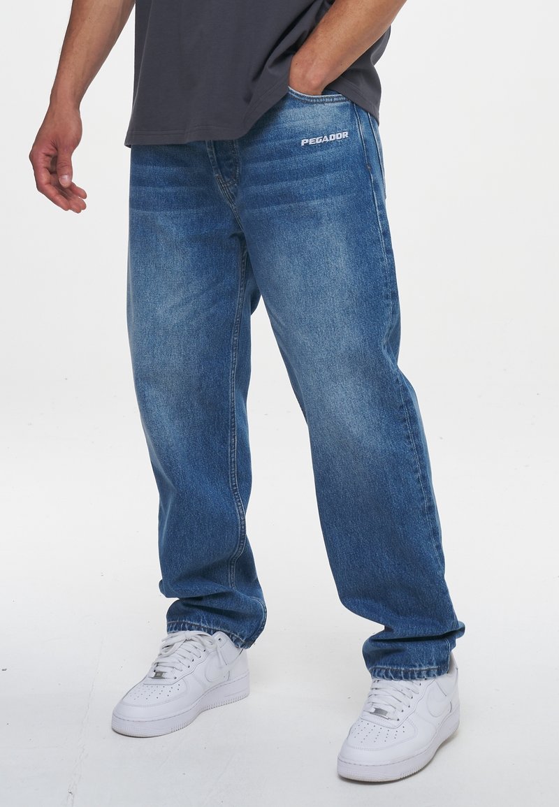 Man med löst sittande blå jeans med "PEGADOR" logotyp på fickan och vita Nike-sneakers, står med en hand i fickan.