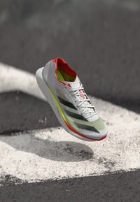Chaussure de course avec un dessus en mesh blanc et gris, accents rouges, forme allongée, design léger et une semelle rembourrée de couleur vive.