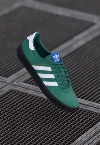 Sapatilhas verdes de camurça com riscas brancas, sola de borracha texturizada preta,logótipo azul e branco na lingueta, e "MONTREAL" escrito em dourado.