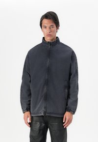 Casaco de ganga preto com gola alta, fecho por zip na frente, bolsos laterais e punhos elásticos, apresentando um ajuste ligeiramente solto e uma textura desgastada.