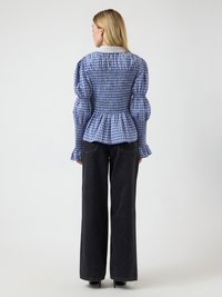 Γυναίκα που κοιτάζει προς τα πίσω, φορώντας μπλε πουκάμισο με ρεβέρ καρό και φουσκωτά μανίκια, καθώς και μαύρο τζιν παντελόνι με φαρδιά πόδια και ψηλοτάκουνα.