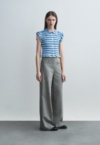 Γυναίκα όρθια, φοράει μπλούζα με κοντά μανίκια, μπλε και λευκό καρό, με βολάν στα μανίκια και γκρι παντελόνι με φαρδιά πόδια, συνοδευόμενο από μαύρα παπούτσια.