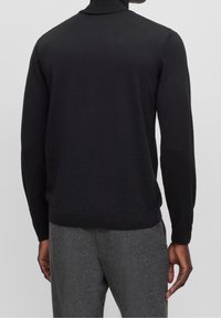 Man som bär en svart långärmad polotröja och gråa byxor, sedd bakifrån mot en enkel ljus bakgrund.