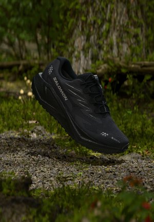 Scarpa da corsa Salomon nera con Gore-Tex su terreno roccioso e muschioso nella foresta, che sembra sospesa in aria come se fosse in movimento.