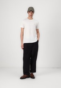 Tricou alb cu mânecă scurtă și un mic logo, pantaloni negri lejeri și loferi maro închis. Modelul poartă o șapcă verde deschis.