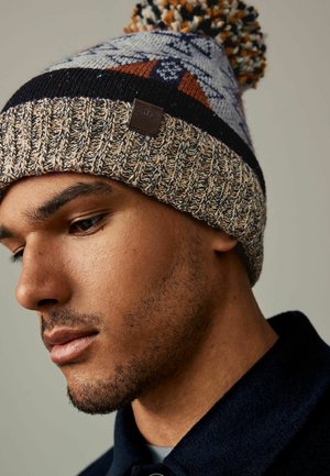 Strikket vinterhue i beige med et mønstret blåt og brunt design. Har en pom-pom på toppen og et struktureret ribkant.