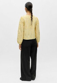 Blouse jaune brodée avec manches bouffantes, dotée d'une ouverture en forme de trou de serrure dans le dos, accompagnée d'un pantalon noir ample. Motif floral texturé sur l'ensemble.