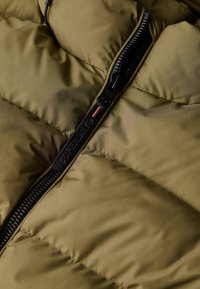 Olijfgroene pufferjack met een gequilte design en een zwarte rits met een logo, heeft een gladde textuur en stevige hardware.