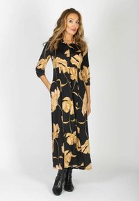 Czarna długa suknia w duży beżowy kwiatowy wzór, z buttonowym dekoltem i rękawami o długości trzech czwartek. Gładki materiał elegancko się układa.