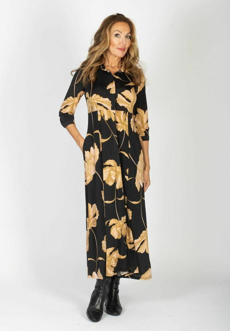 Czarna długa suknia w duży beżowy kwiatowy wzór, z buttonowym dekoltem i rękawami o długości trzech czwartek. Gładki materiał elegancko się układa.