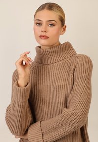 Maglione beige lavorato a maglia con colletto alto, texture a coste e maniche larghe. Il tessuto appare morbido e caldo.