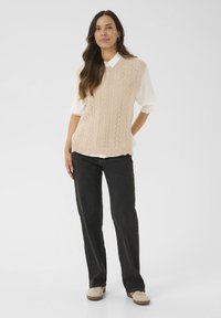 Femme portant un gilet en tricot sans manches beige sur une blouse blanche à manches bouffantes, un jean noir droit et des chaussures beiges, se tenant avec une main derrière le dos.