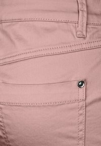 Svetle růžové kalhoty s hladkou texturou látky, zdobené viditelným švem a malým tmavým kovovým doplňkem na kapse.