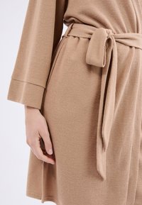Robe beige con texture, dotato di un girovita annodato, maniche lunghe e una vestibilità ampia, caratterizzato da un dettagliato motivo tessuto sul tessuto.
