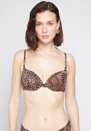 Mujer con cabello corto castaño, con pendientes de aro dorados y un sujetador de estampado de leopardo, mirando hacia adelante sobre un fondo liso.