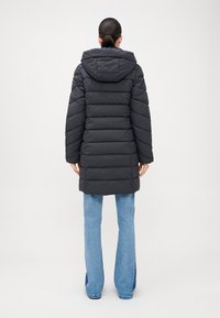 Juodas pūkuotas parka paltas su kapišonu, šoninėmis kišenėmis ir horizontaliu siuvimu. Vilkimas ant mėlynų plačių džinsų.
