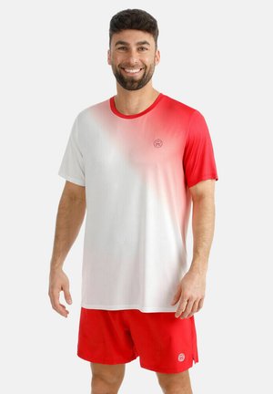 Lächelnder Mann mit dunklem Haar und Bart, der ein rot-weiß verlaufendes Sportshirt und rote Shorts trägt, stehend vor einem weißen Hintergrund.