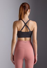 Crna sportska brassiere s ukriženim trakama i logotipom, u kombinaciji s visokim, svijetlo ružičastim tajicama koje imaju bešavni dizajn.
