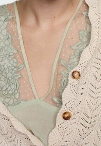 Světle béžový pletený cardigan s vzorem listů, doplněný dvěma hnědými knoflíky, přeložený přes mátově zelený top s květinovou výšivkou.