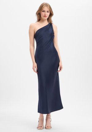 Frau steht, trägt ein ärmelloses, marineblaues Satin-Ein-Schulter-Langkleid und beigen High-Heel-Sandalen auf weißem Hintergrund.