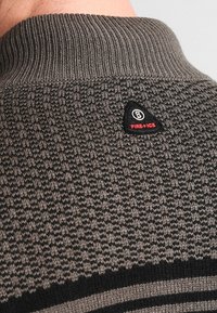 Zbliżenie na szary sweter z dzianiny, na szyi osoby, z małą czarną łatą w kształcie trójkąta oznaczoną "FIRE + ICE".