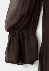 Detalle de blusa marrón transparente, con un tejido texturizado, puños fruncidos y mangas fluidas, con una calidad semitransparente sutil.
