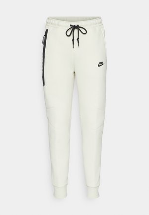 Pantaloni jogger Nike in crema chiaro, con logo nero, coulisse regolabile, tasche laterali con zip e design a gamba affusolata.