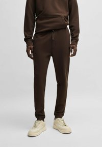 Bruine sweatshirt met een voorzak en bijpassende bruine joggers. De stof lijkt zacht en gestructureerd. Gecombineerd met lichte sneakers.