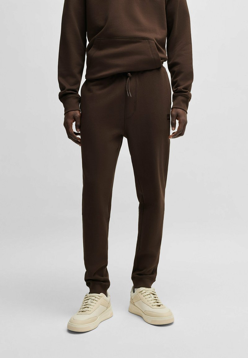 Bruine sweatshirt met een voorzak en bijpassende bruine joggers. De stof lijkt zacht en gestructureerd. Gecombineerd met lichte sneakers.