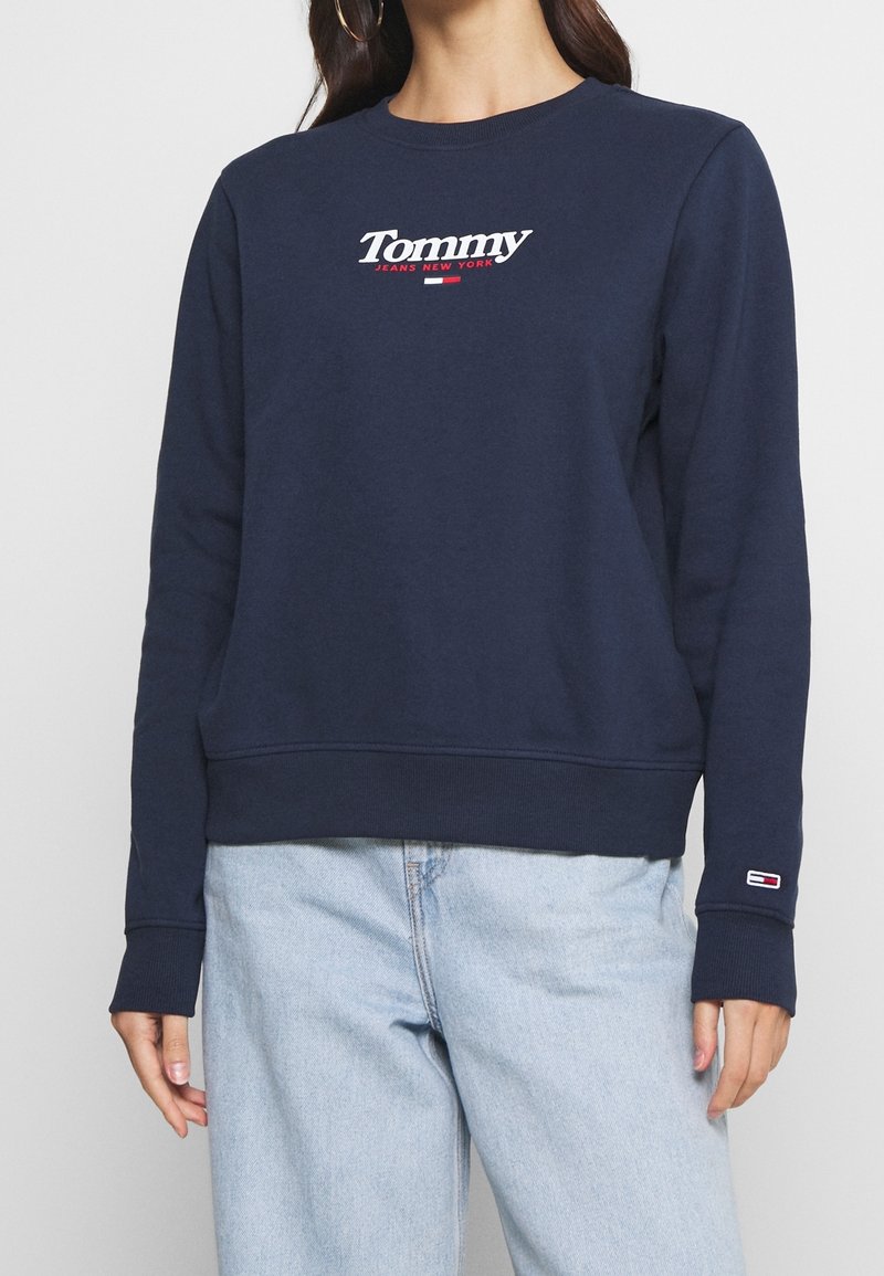 Femme portant un sweat-shirt Tommy Jeans bleu marine avec logo sur la poitrine et les poignets, associé à un jean bleu clair, sur fond uni.