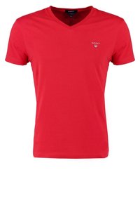 T-shirt en coton rouge avec col en V, manches courtes et petit logo blanc sur la poitrine à gauche. Texture lisse et coupe standard.