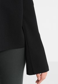 Black knit pullover with a loose fit and wide sleeves, featuring side slits. Paired with dark green pants. Smooth texture.