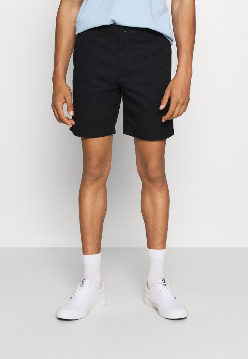 Homme portant un short noir, des chaussettes blanches et des baskets blanches, debout devant un fond clair uni.