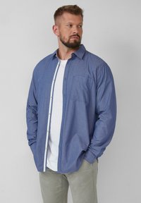 Niebieska longsleeve z białym wzorem w kropki, zapinana na guziki z jedną kieszonką na piersi i kołnierzykiem, noszona na białej koszulce.