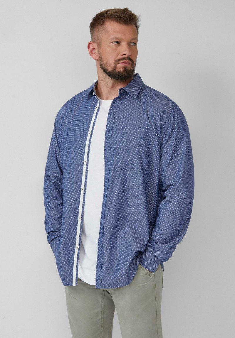 Niebieska longsleeve z białym wzorem w kropki, zapinana na guziki z jedną kieszonką na piersi i kołnierzykiem, noszona na białej koszulce.