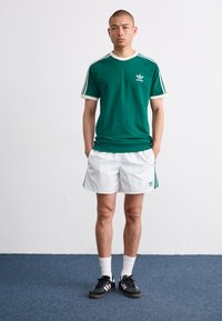 Zöld pamut póló fehér részletekkel és Adidas logóval, fehér sorttal, zöld oldalsávokkal, valamint fekete sportcipővel párosítva.