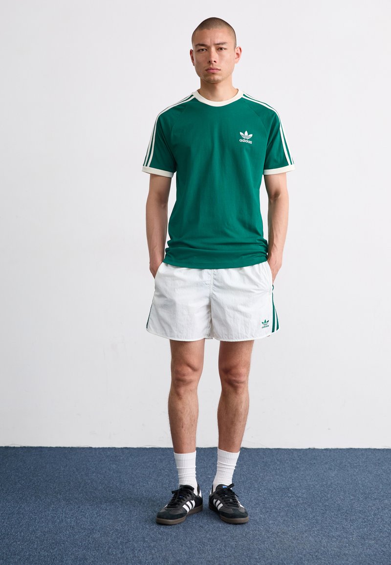 Camiseta de algodón verde con acentos blancos y logo de Adidas, combinada con pantalones cortos blancos con franjas laterales verdes y zapatillas negras.