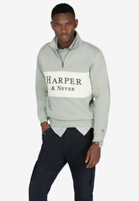 Hellgrauer Reißverschluss-Sweatshirt mit einem weißen Brustpanel, auf dem "HARPER & Neyer" in schwarzer Schrift steht. Kombiniert mit dunklen Cargo-Hosen und einem gestreiften Hemd.