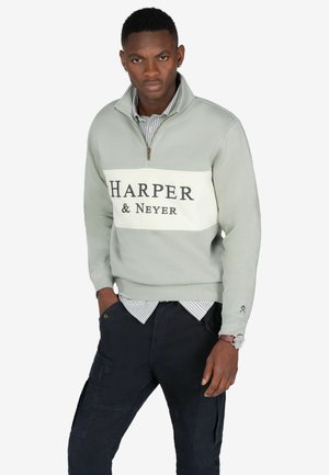 Sudadera de cremallera gris claro con un panel en el pecho blanco que lleva el texto "HARPER & Neyer" en negro. Combinada con pantalones cargo oscuros y una camiseta a rayas.