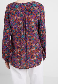 Blusa floreale a maniche lunghe con sfondo arancione, caratterizzata da fiori viola e verde aqua. Tessuto morbido, vestibilità rilassata e fondo arrotondato.