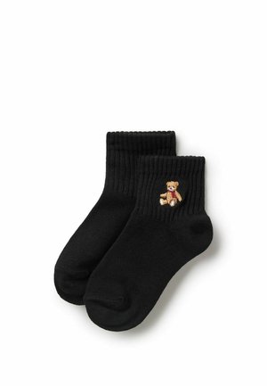 Schwarze gerippte Knöchelsocken mit einem kleinen braunen bestickten Teddybär, der einen roten Schal trägt, auf der Außenseite einer Socke.