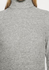 Maglione grigio a collo alto lavorato a maglia con una texture a costine, che presenta una vestibilità aderente e un colletto allungato, mostrando un aspetto morbido e accogliente.