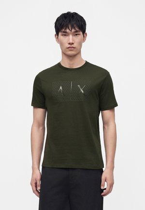 Homme portant un t-shirt vert foncé à manches courtes avec un logo A|X en relief et un pantalon noir, debout devant un fond clair uni.