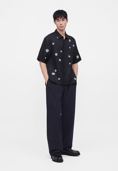 Un homme se tient debout, les mains dans les poches, portant une chemise noire à manches courtes à boutons avec des imprimés floraux blancs, un pantalon ample foncé et des chaussures noires.