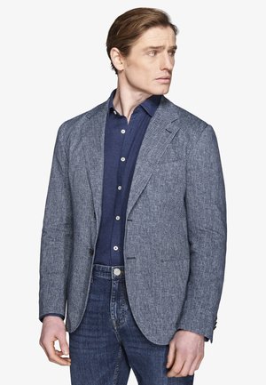 Mann trägt ein graues Fischgrätenmuster-Sakko über einem dunkelblauen Hemd mit Knöpfen und blauen Jeans, schaut nach rechts.