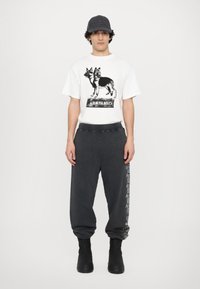 Weißes T-Shirt mit einem schwarzen Grafikdesign von Hunden, kombiniert mit dunkelgrauen Sweatpants, die seitlich mit Text bedruckt sind, und schwarzen Stiefeln.